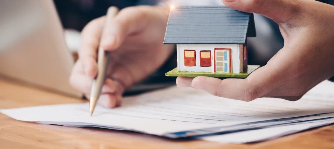 Um homem analisa um contrato de doação de imóvel antes de assinar. Ele segura a miniatura de uma casa, além de papel e caneta.
