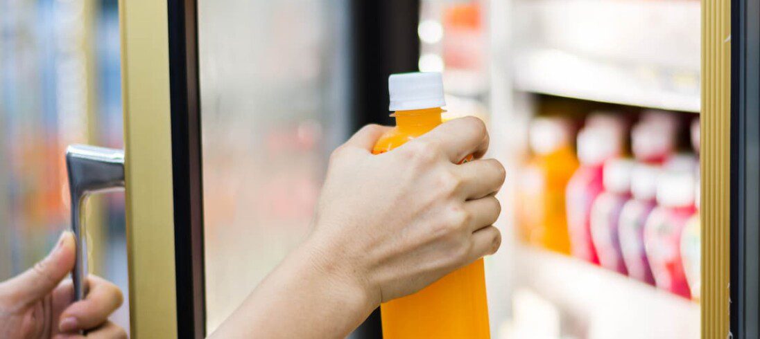 Um pessoa pega um suco de laranja em uma geladeira de mercadinho em condomínio.