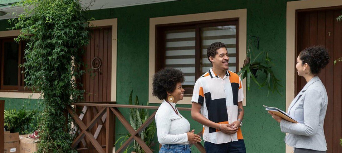 Foto que ilustra matéria sobre casa de vila mostra um casal sorridente conversando com uma corretora de imóveis em frente a uma casa verde