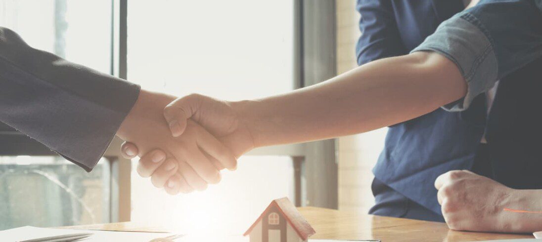 Duas pessoas fazem acordo sobre de repasse imobiliário. Elas apertam as mãos ao lado de um contrato e a miniatura de uma casa.