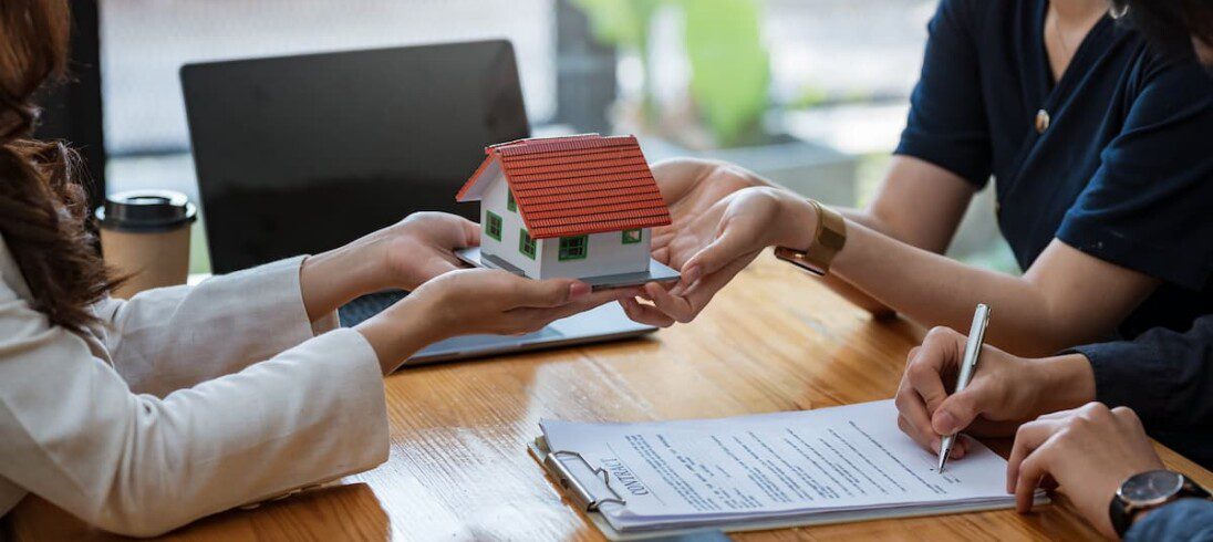 Duas pessoas assinam um contrato de compra conjunta de imóvel. Uma mulher, provavelmente de uma imobiliária, passa a miniatura de uma casa para um dos compradores.
