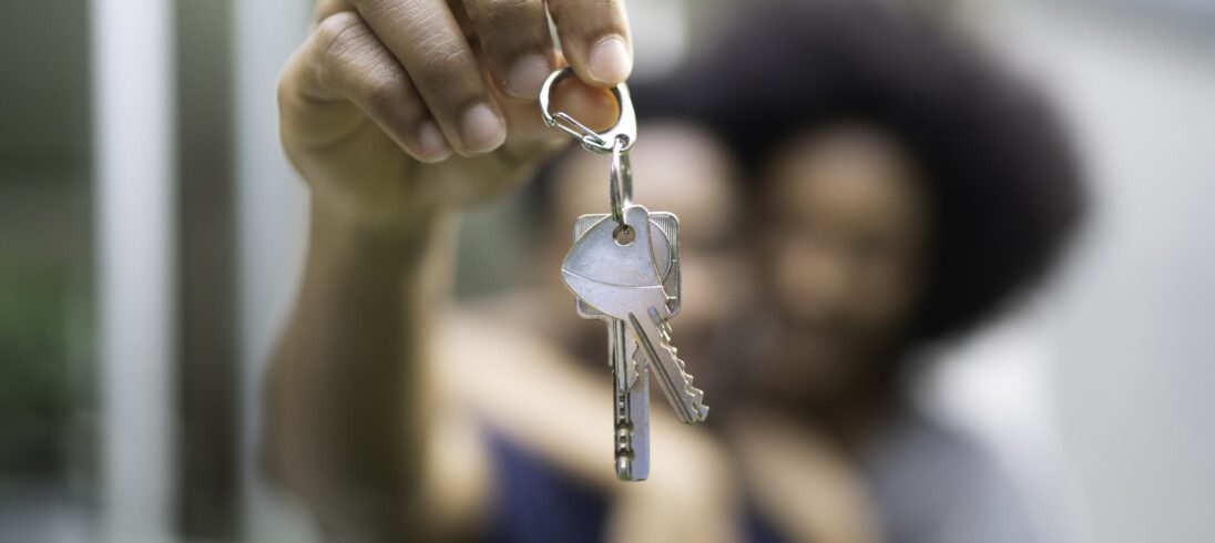 Imagem que ilustra matéria sobre comprar imóvel agora ou esperar 2023 mostra um casal ao fundo desfocado segurando chaves na mão