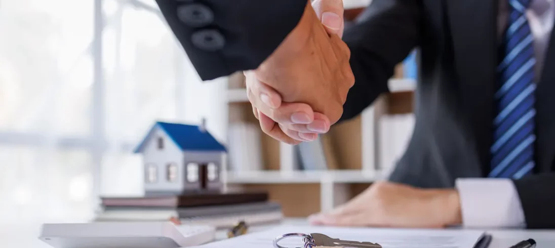 Dois homens apertam as mãos fechando um contrato. Há uma chave em cima do contrato e a miniatura de uma casa.