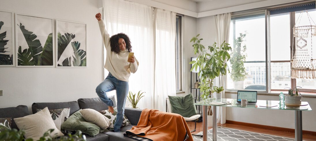 Imagem de uma jovem feliz e animada escutando música em cima do sofá, com um dos braços para cima, em um apartamento com estilo jovial, com uma janela à direita e plantas à esquerda, para decorar matéria sobre apartamento para solteiro