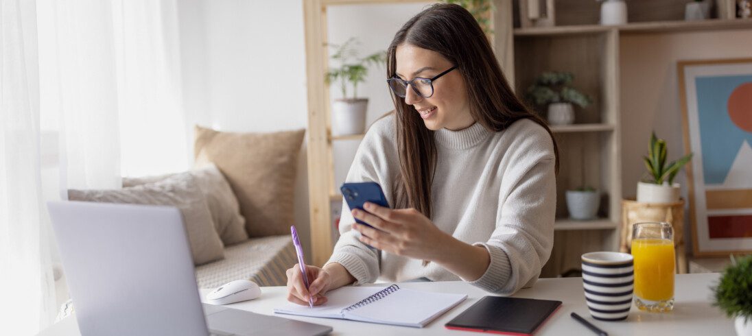 Imagem de uma mulher trabalhando em casa tomando notas no caderno e no celular para ilustrar matéria sobre como economizar dinheiro para comprar um apartamento