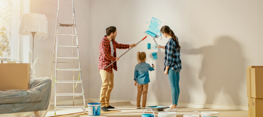 Foto de uma mulher, um homem e uma criança pequena pintando a parede de casa com um rolo pintura mergulhado em tinta azul para ilustrar matéria sobre como economizar na reforma da casa