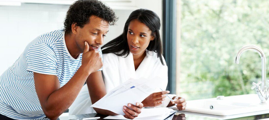 Um casal de homem e mulher negros olham um documento em cima de uma bancada na cozinha.