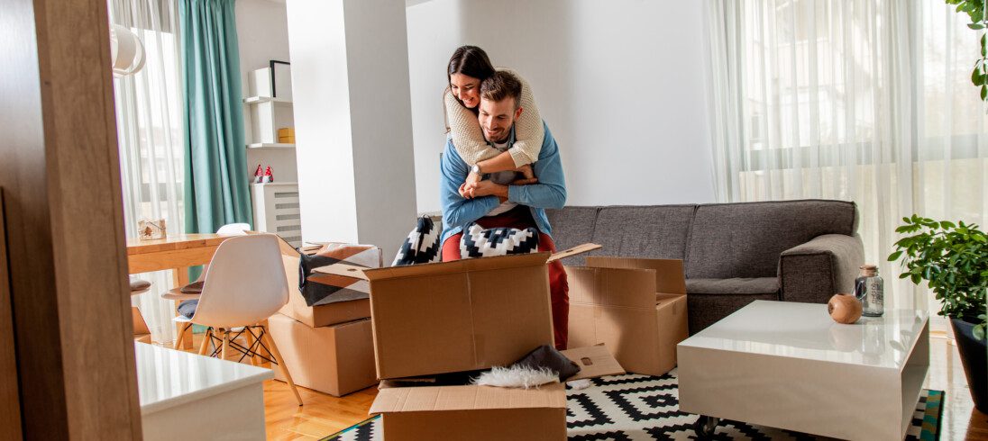 Imagem de um casal composto por uma mulher e um homem abrindo uma caixa em um apartamento mobiliado para ilustrar matéria sobre as vantagens de morar em apartamento