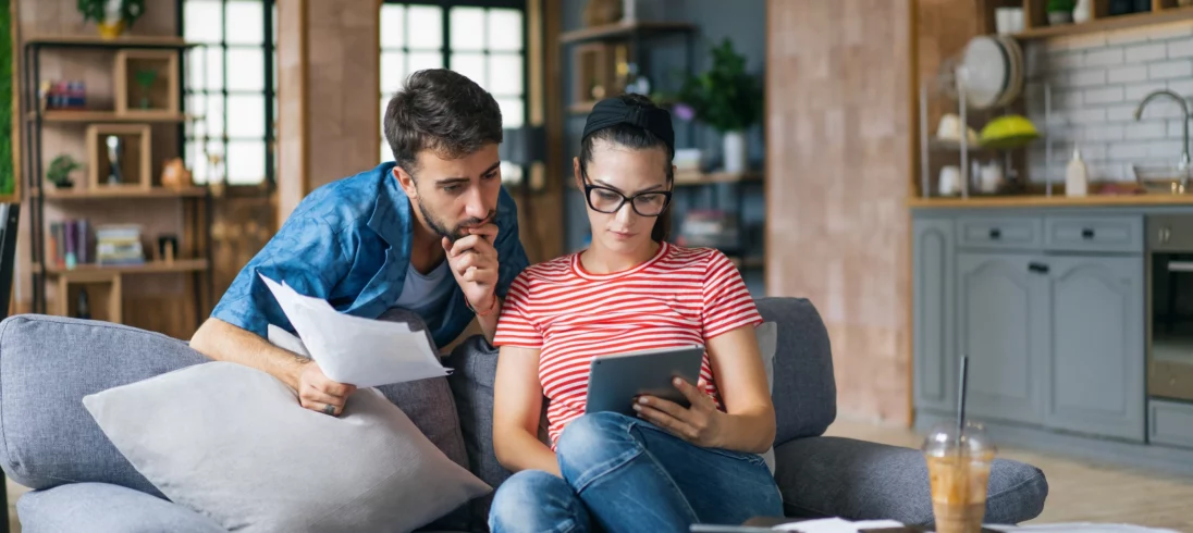 Foto de um casal olhando para um tablet com papéis na mão no sofá da sala de uma casa para ilustrar matéria sobre como comprovar posse de imóvel