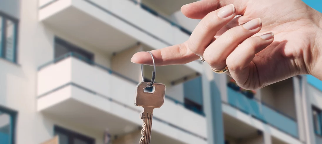Imagem de uma mão feminina segurando a chave de um imóvel em frente a um prédio residencial para ilustrar matéria sobre como escolher um bom apartamento