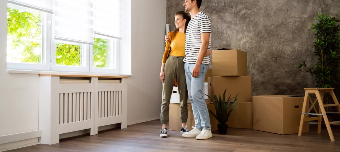 Imagem de um casal formado por uma mulher e um homem abraçados no ambiente interno de uma casa, sorrindo um para o outro, para ilustrar matéria sobre como comprar uma casa