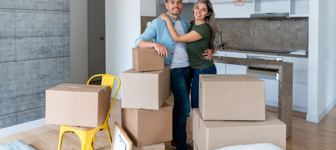 Imagem de um casal, composto por um homem e uma mulher, felizes em um apartamento. Eles estão parados e sorrindo atrás de algumas caixas de papelão para ilustrar matéria sobre tamanho de apartamento