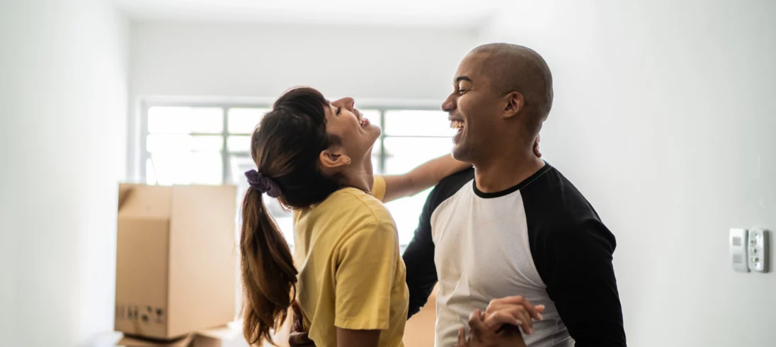 Imagem de um casal composto por um homem e uma mulher dançando com expressão de felicidade no rosto em sua casa nova para ilustrar matéria sobre como financiar 100% de um imóvel