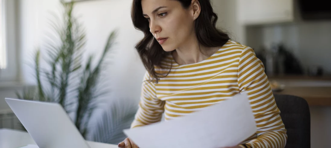 Imagem de uma mulher jovem e branca com uma expressão séria sentada olhando para a tela de um computador com alguns papéis na mão para ilustrar a consulta de imóveis por CPF grátis