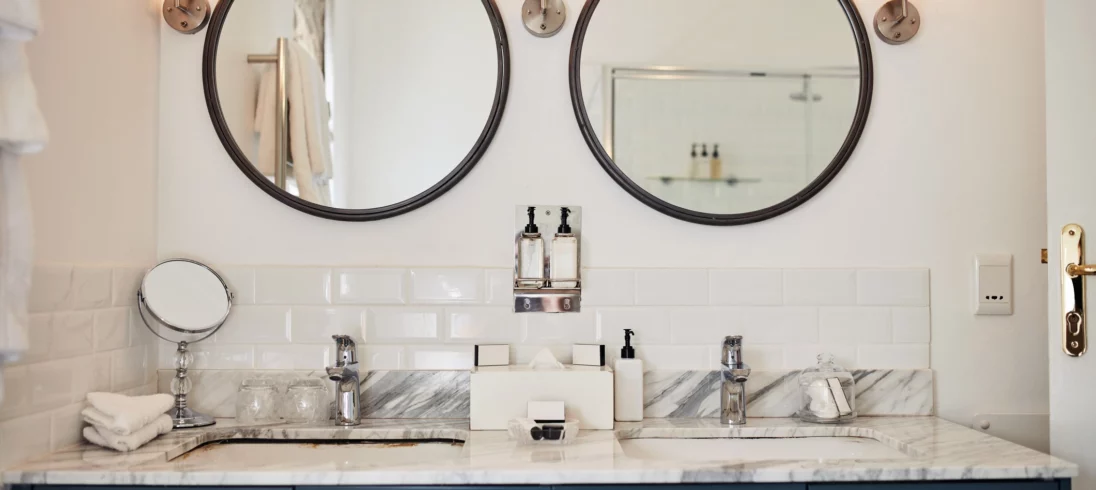 Imagem de um banheiro de demi-suíte com parede clara, dois espelhos redondos com moldura preta posicionado lado a lado, com luminárias entre eles. Um armário azul, com duas cubas e torneiras. Na bancada das pias estão posicionados produtos de higiene pessoal, toalhas e um espelho de maquiagem