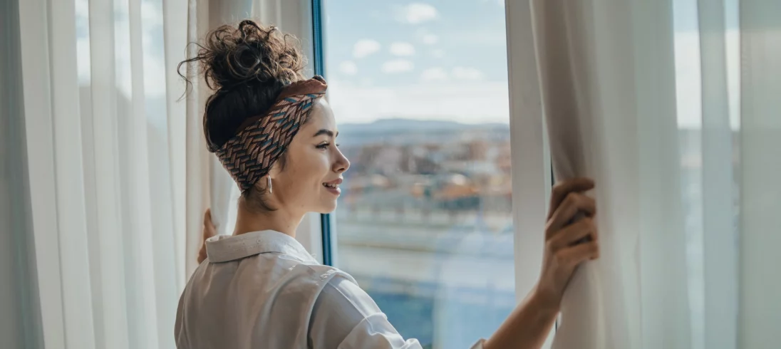 Imagem de uma mulher jovem e sorridente abrindo as cortinas de um apartamento e olhando para o horizonte para ilustrar matéria sobre quanto custa um apartamento