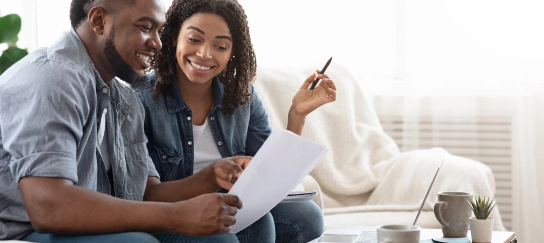 Imagem de um casal de jovens negros composto por um homem e uma mulher sorridentes sentados no sofá de uma casa olhando para um documento para ilustrar matéria sobre termo de quitação de imóvel