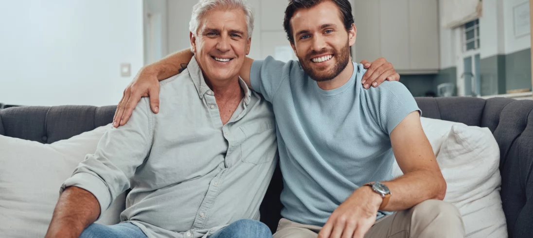 Imagem de um homem idoso com seu filho adulto, sentados um ao lado do outro em um sofá com expressão sorridente para ilustrar matéria sobre venda de imóvel para filho