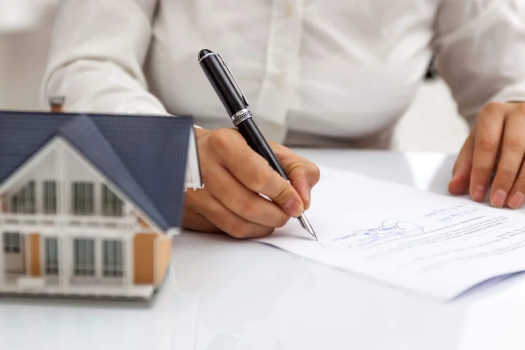 Imagem de uma pessoa assinando um documento em cima de uma mesa ao lado de uma miniatura de uma casa para ilustrar matéria sobre contrato de aluguel com fiador
