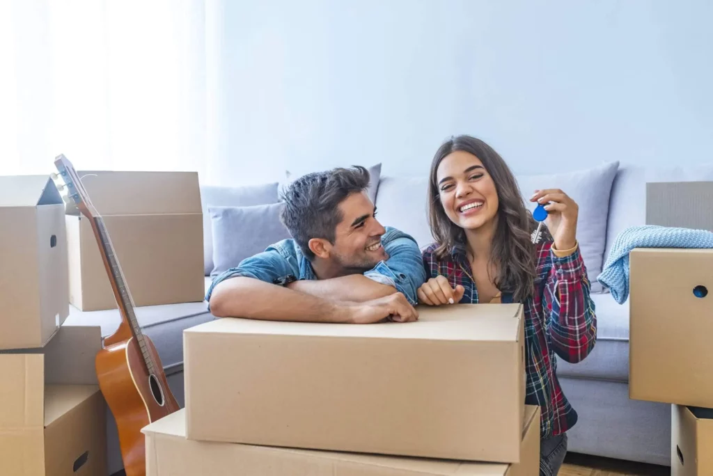 Imagem de um casal composto por um homem e uma mulher em uma sala de estar com várias caixas de papelão. Eles estão sorrindo e segurando a chave do imóvel para ilustrar matéria sobre o que é fiador de aluguel
