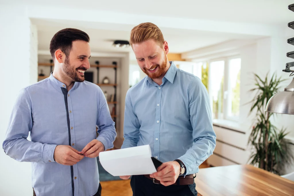 Imagem de dois homens em pé em uma sala de estar de uma casa sorrindo e segurando documentos para ilustrar matéria sobre o termo de entrega de chaves.