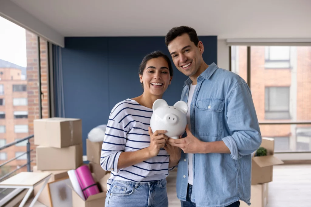 Imagem de um casal feliz composto por um homem e uma mulher em uma casa com várias caixas de papelão e segurando um cofre em formato de porquinho para ilustrar matéria sobre qual o score ideal para financiamento de imóvel