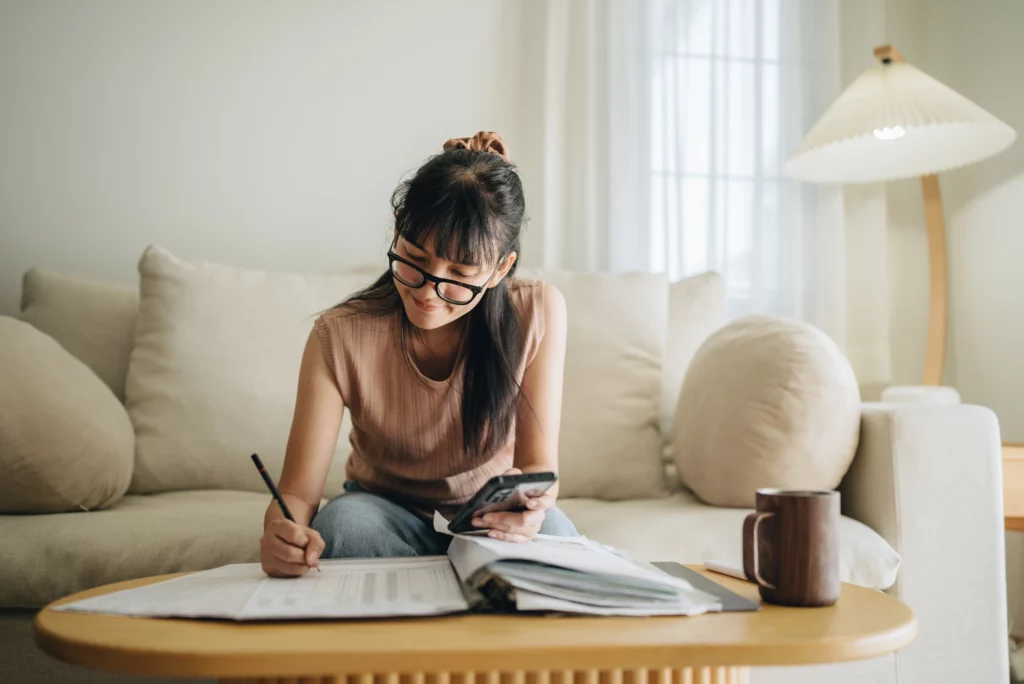 Imagem de uma mulher planejando orçamento financeiro com um celular, um caderno e um lápis para ilustrar matéria sobre quanto custa morar sozinho