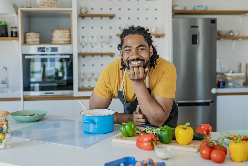 Imagem de um homem jovem na cozinha de casa encostado na bancada com a mão no queixo sorrindo em frente a uma panela azul, utensílios de cozinha e legumes diversos para ilustrar matéria sobre as despesas de morar sozinho
