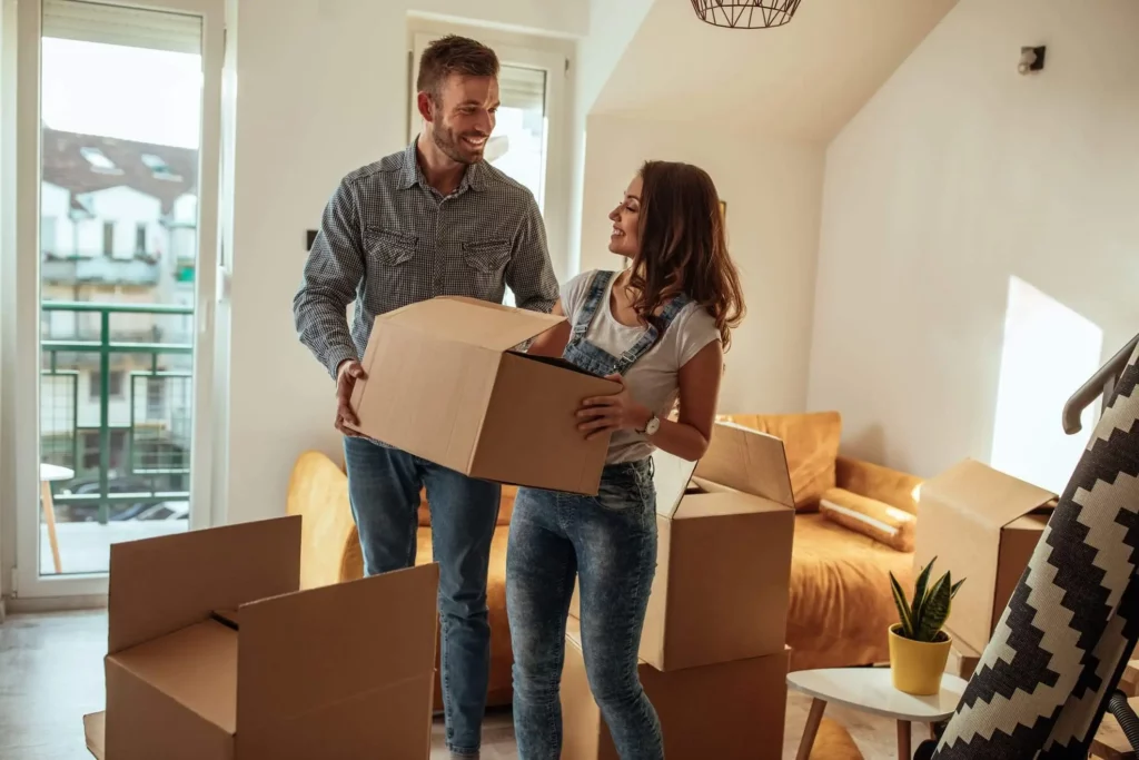 Imagem de um casal composto por um homem e uma mulher transportando uma caixa de papelão em uma sala de estar para ilustrar matéria sobre quanto custa um fiador de aluguel