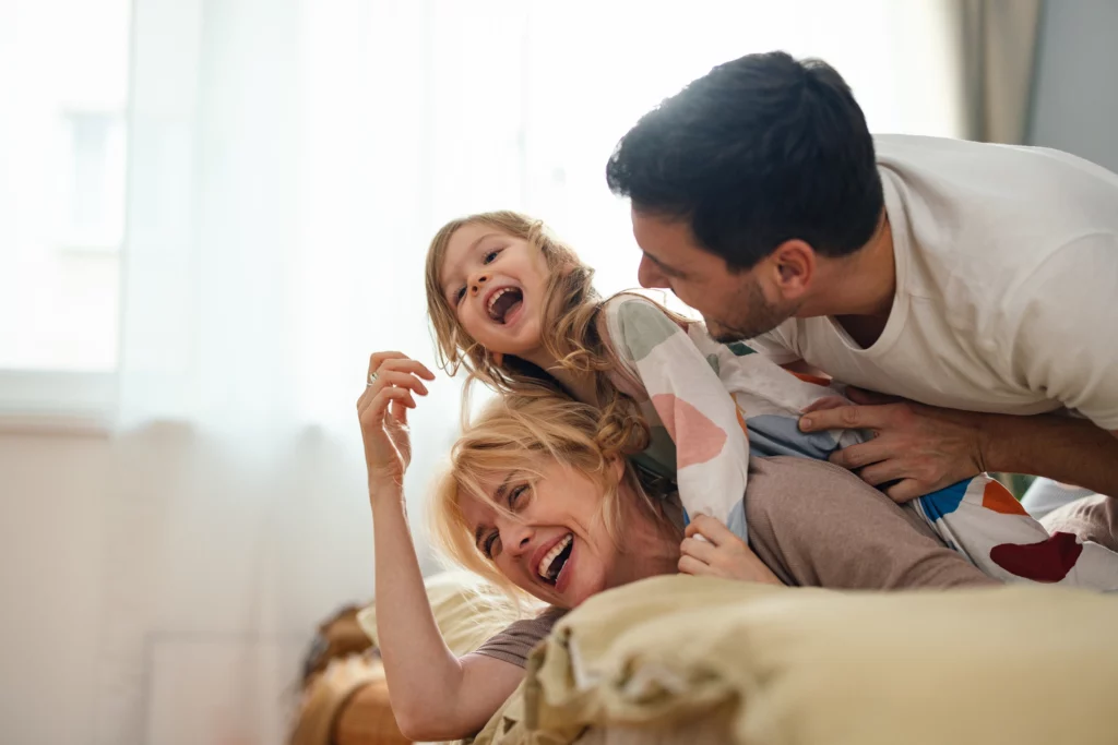 Imagem de uma família feliz composto por um homem, uma mulher e uma criança em cima da cama sorrindo para ilustrar matéria sobre quanto de score precisa para financiar um imóvel