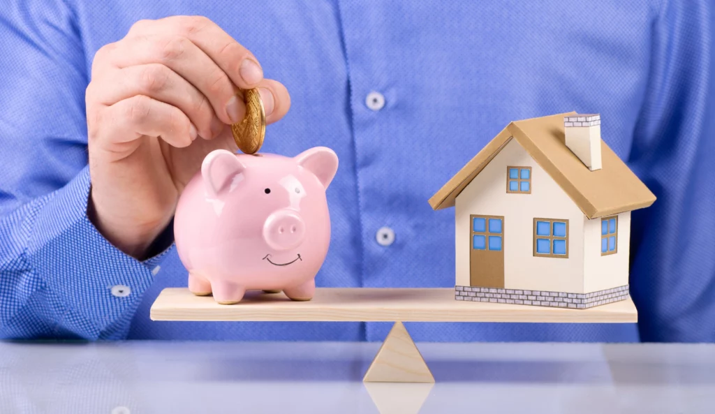  Imagem de uma pessoa ao fundo colocando uma moeda em um cofre em formato de porquinho que está do lado esquerdo de uma balança equilibrada com uma casa de madeira do lado direito para ilustrar matéria sobre quanto preciso para morar sozinho