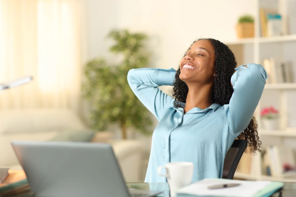 Imagem de uma mulher negra em casa sentada em uma mesa feliz em frente ao computador para ilustrar matéria sobre como alugar imóvel com score baixo