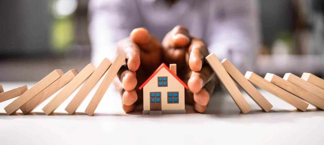 Imagem de um dominó caindo sobre a miniatura de uma casa e sendo barrado pelas mãos de uma pessoa para ilustrar matéria sobre seguro prestamista