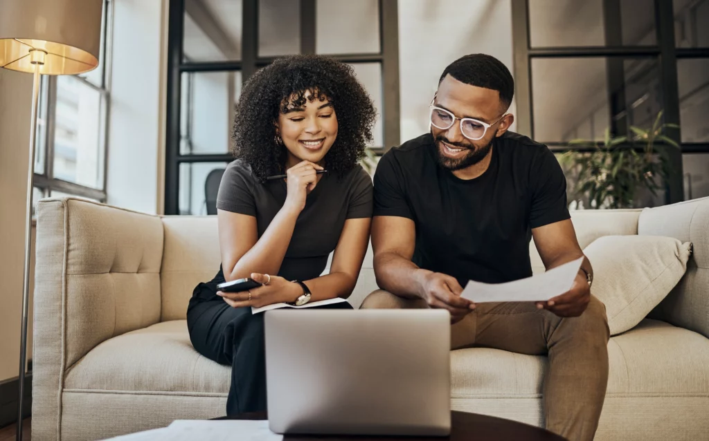 Imagem de um casal negro composto por um homem e uma mulher sentados no sofá em frente a um computador analisando informações para ilustrar matéria sobre seguro prestamista