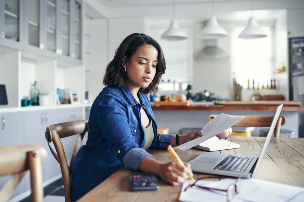 Imagem de uma mulher sentada na mesa da cozinha de uma casa usando o computador para ilustrar matéria sobre as vantagens de cobertura para um financiamento imobiliário