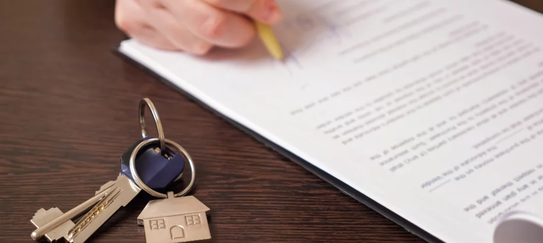 Imagem de uma pessoa assinando um papel em cima de uma mesa onde há um molho de chaves para ilustrar matéria sobre o termo de entrega de chaves