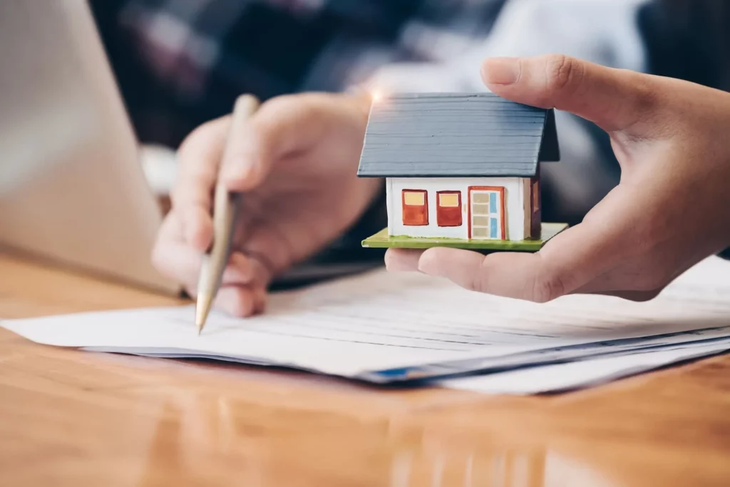 Imagem de uma pessoa segurando uma miniatura de uma casa enquanto assina um documento em uma mesa para ilustrar matéria sobre as vantagens de morar em um loteamento
