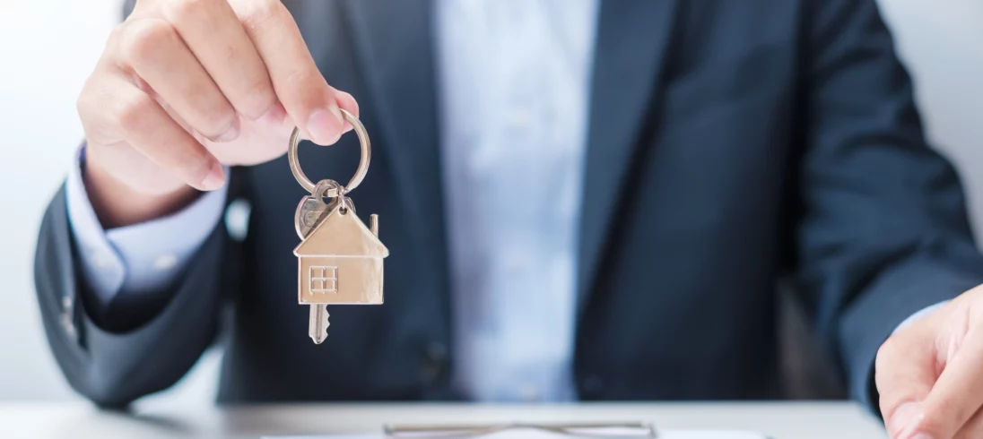 Imagem de um homem de terno segurando uma chave com chaveiro de casa enquanto apresenta uma prancheta com documento e caneta para ilustrar matéria sobre despachante imobiliário