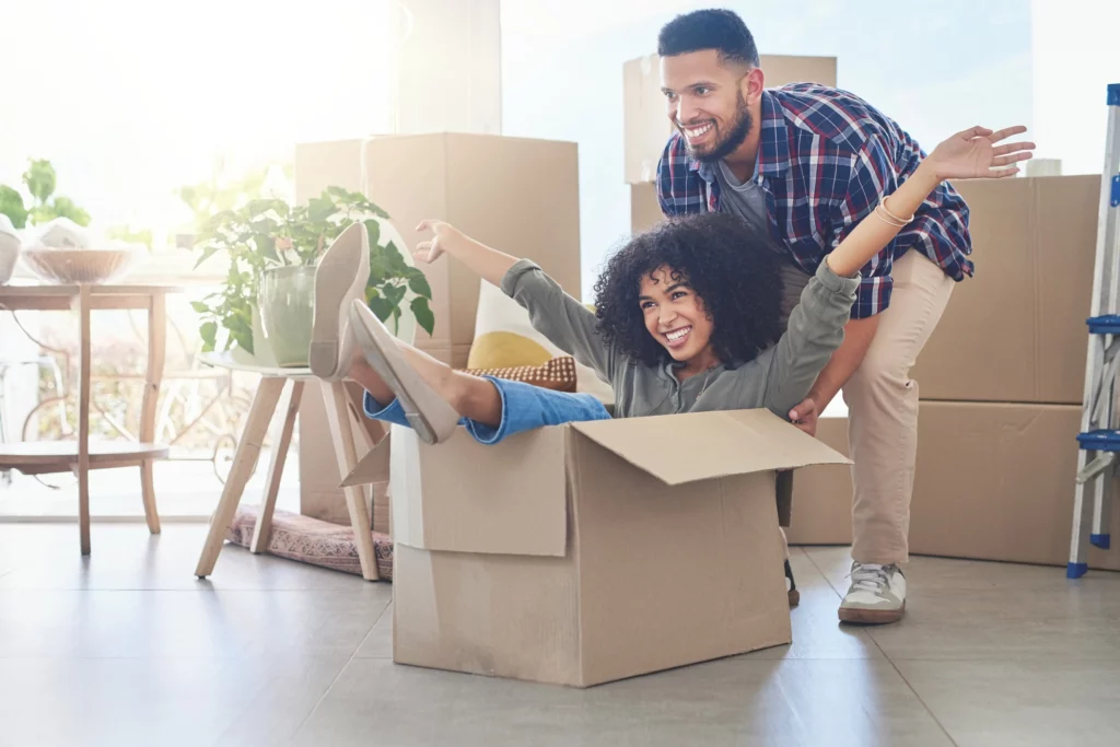 Imagem de casal jovem onde o homem empurra uma mulher em uma caixa vazia em uma casa nova para ilustrar matéria sobre por que contratar um despachante imobiliário
Título: por que contratar um despachante imobiliário