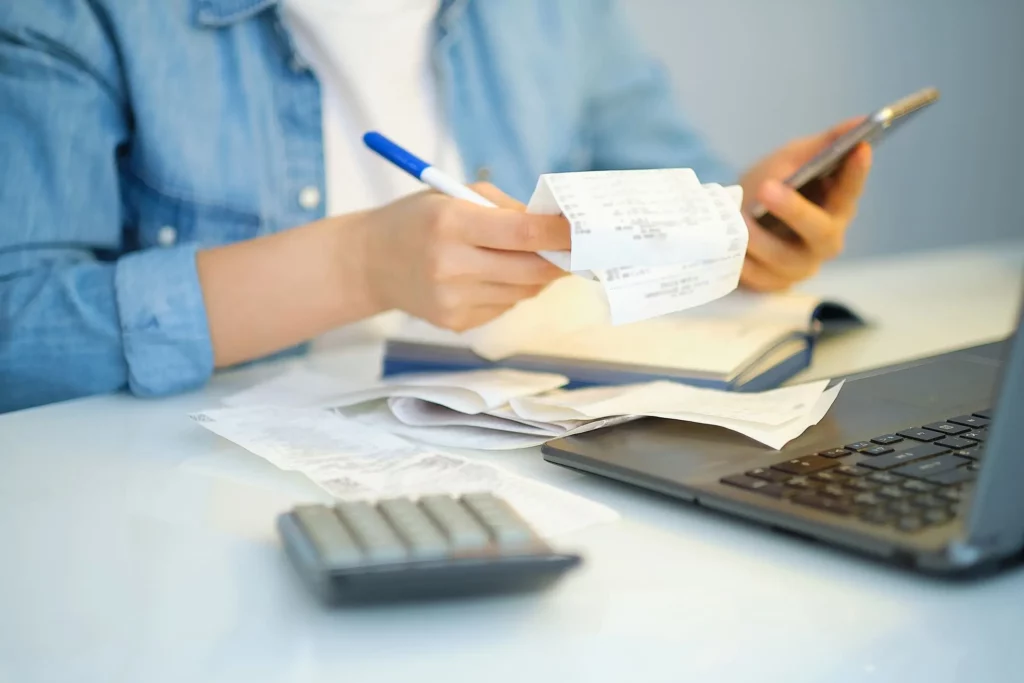 Imagem de uma pessoa sentada em frente a um computador usando o celular e a calculadora para calcular dívidas para ilustrar matéria sobre qual score ideal para financiamento de imóvel