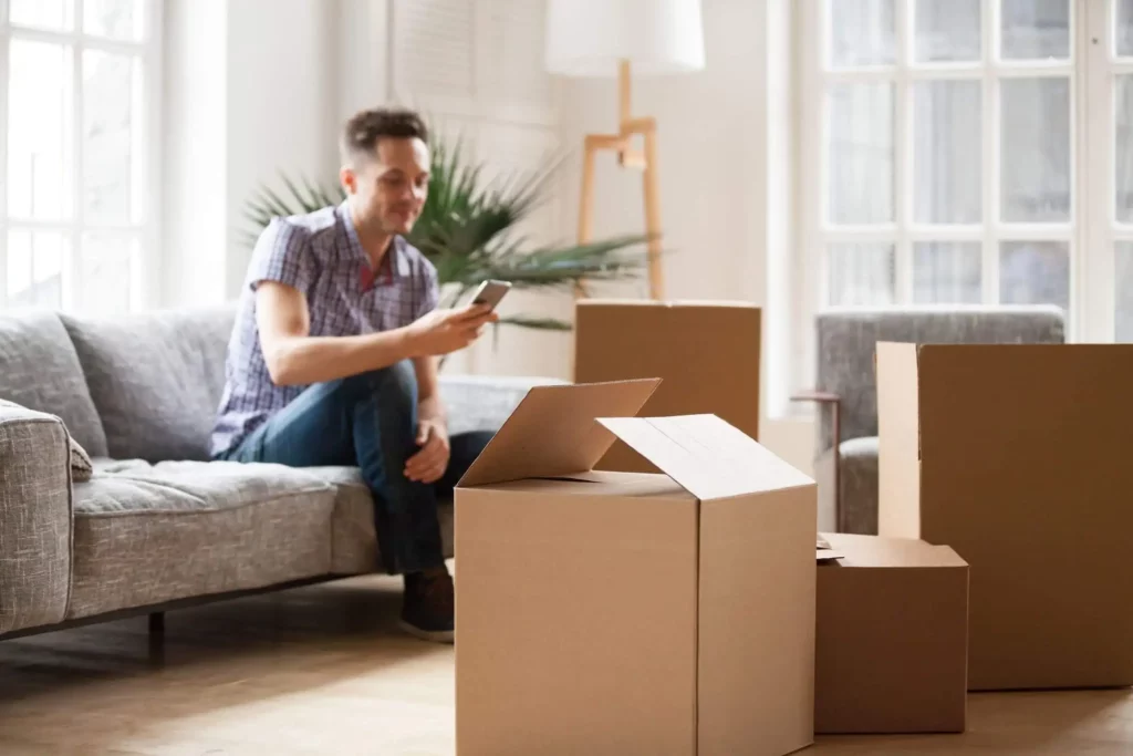 Imagem de um homem sentado no sofá de uma sala com um celular na mão e caixas de papelão espalhadas pelo espaço para ilustrar matéria sobre quanto custa morar em condomínio