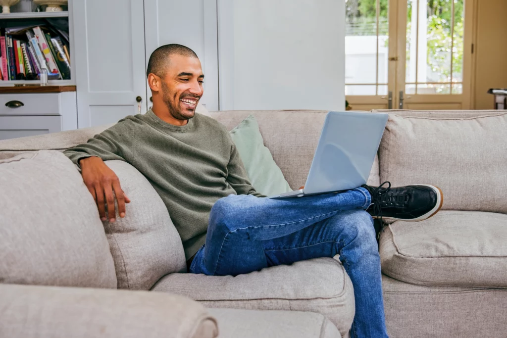 Imagem de um homem sentado no sofá de casa sorrindo com um computador no colo para ilustrar matéria sobre o tamanho ideal de apartamento para 4 pessoas