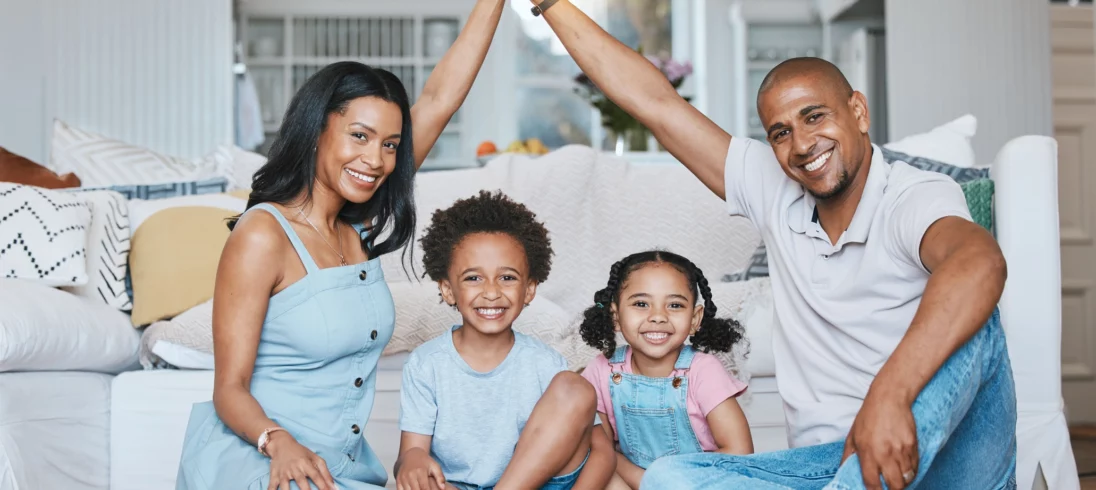 Imagem de uma família composta por um casal e duas crianças negras sentadas no chão da sala sorridentes para ilustrar matéria sobre o tamanho ideal de apartamento para 4 pessoas