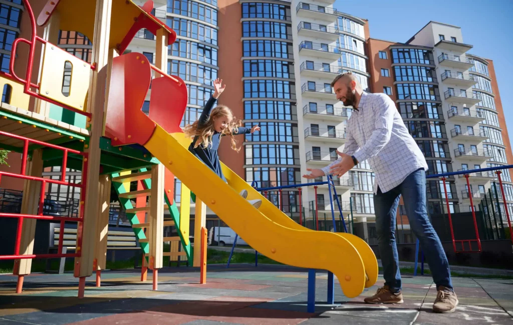 Imagem de uma criança descendo no escorregador de um playground de um condomínio enquanto o pai a espera no final do brinquedo para ilustrar matéria sobre como calcular o valor do condomínio