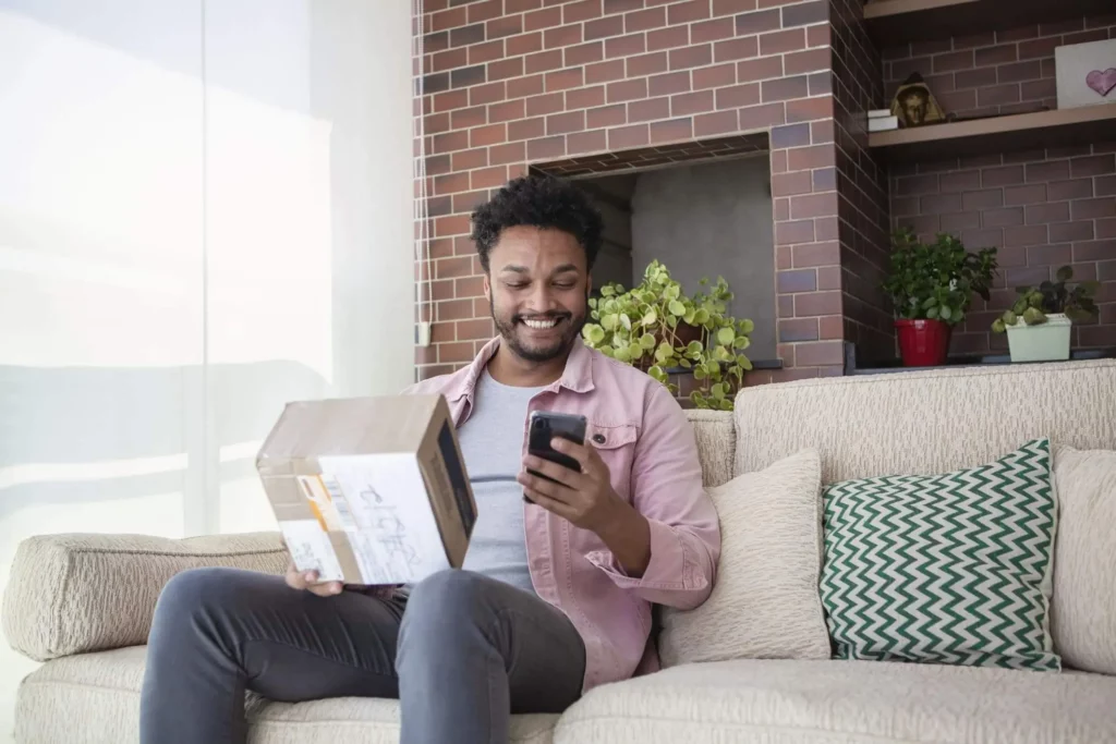Imagem de um homem negro sentado no sofá de casa com uma caixa de papelão e o celular em outra mão para ilustrar matéria sobre como colocar endereço de apartamento para entrega