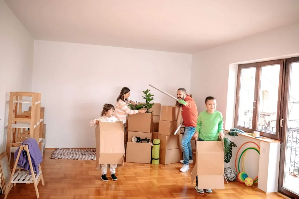 Imagem de uma família composta por um homem, uma mulher e duas crianças carregando caixas de papelão para dentro uma casa para ilustrar matéria sobre habitação de interesse social

