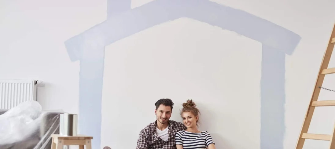 Imagem de um casal composto por um homem e uma mulher sentados no chão de uma casa com alguns itens de obra ao lado e o desenho de uma casa na parede branca para ilustrar matéria sobre habitação de interesse social