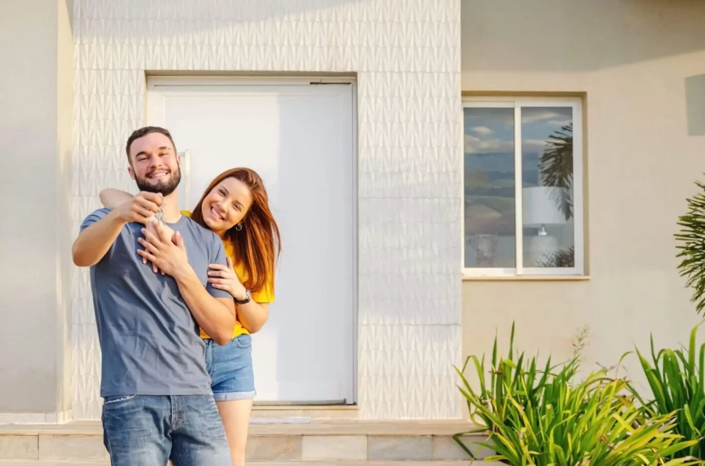 Imagem de um casal composto por um homem e uma mulher abraçados e sorrindo do lado de fora de uma casa para ilustrar matéria sobre a HIS, habitação de interesse social