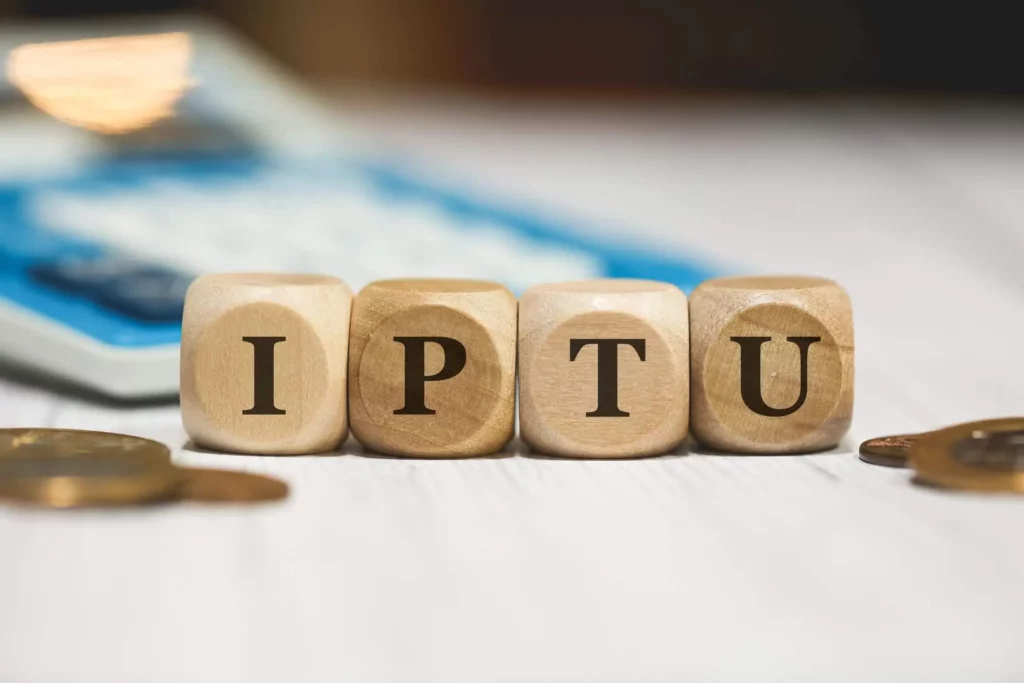 Imagem das letras da sigla “IPTU” escritas em blocos de madeira cercadas por moedas de um real com uma calculadora em segundo plano para ilustrar a matéria sobre o que é o IPTU