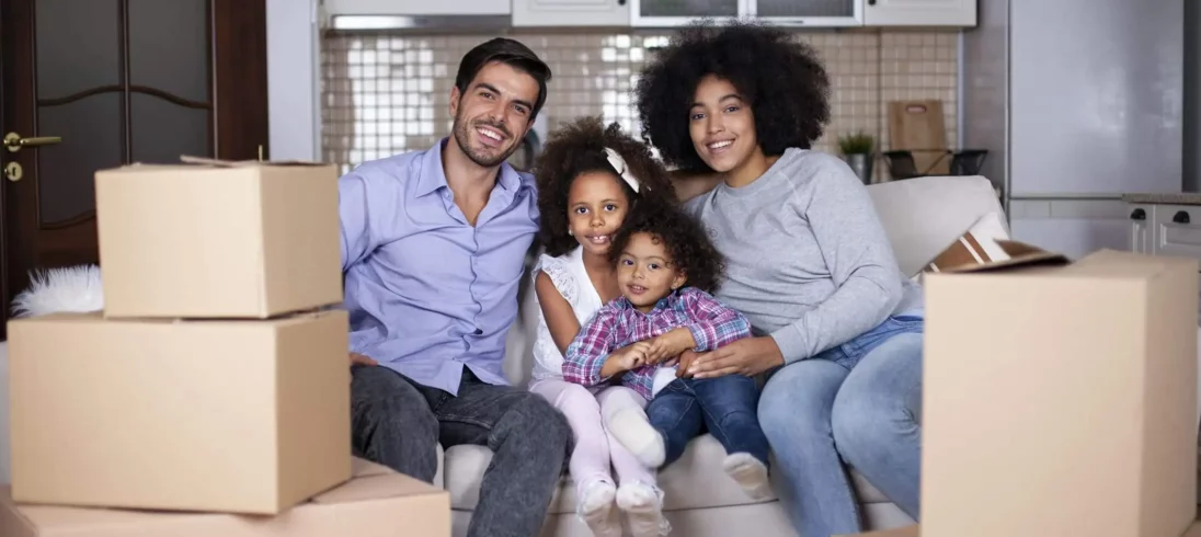 Imagem de uma família dois adultos e duas crianças sorrindo sentadas em um sofá rodeado de caixas de mudança para ilustrar matéria sobre Posso alugar apartamento financiado pela Caixa Minha Casa, Minha Vida